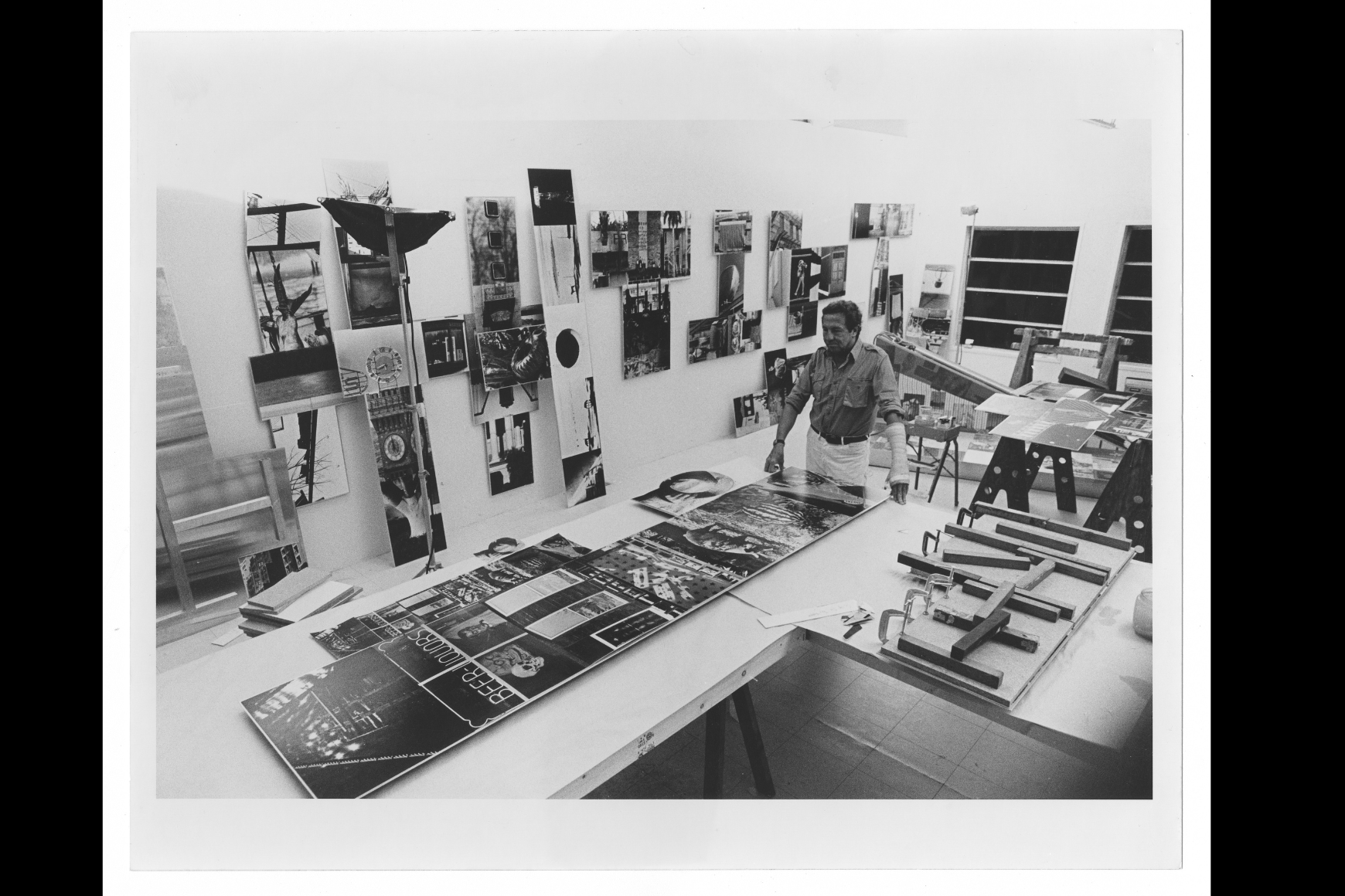 Rauschenberg stands in an art studio, examining large prints on a table surrounded by tools and materials, with artworks and photographs displayed on the walls. 